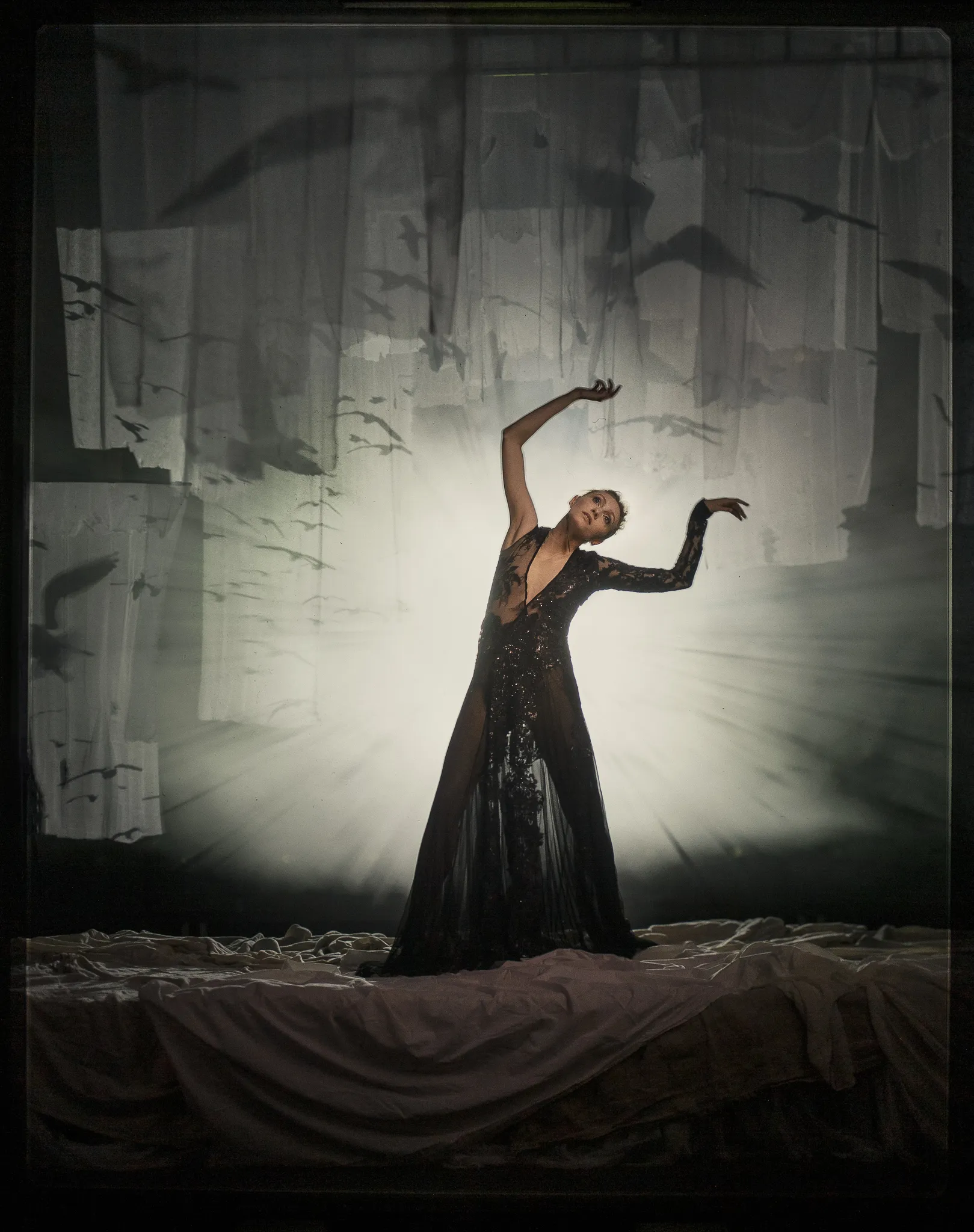 Maria Bolshova as Zarechnaya raises her arms in a sheer black gown against bright backlight.