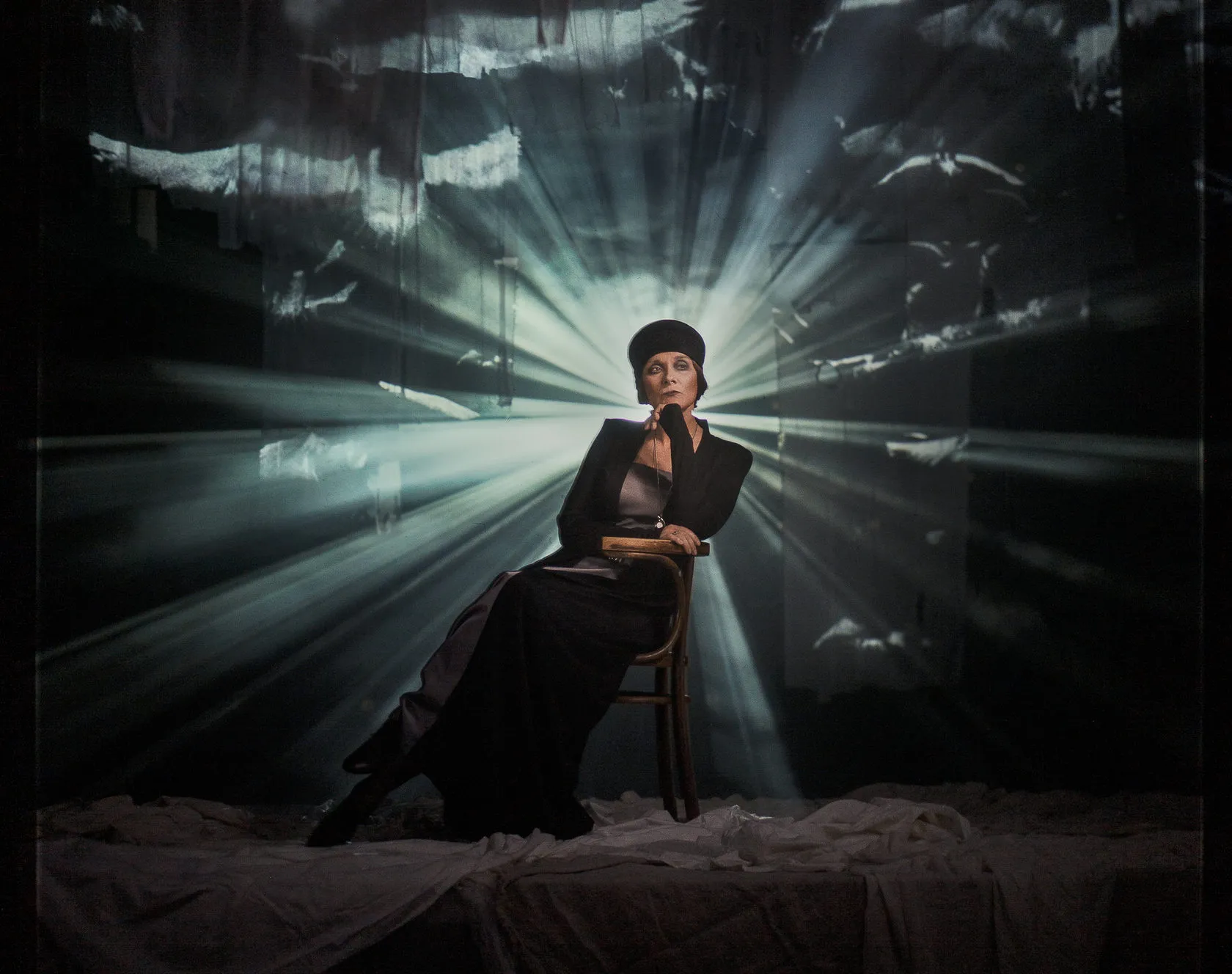 An actress in a black cap sits on a chair in a burst of light and projected gulls.