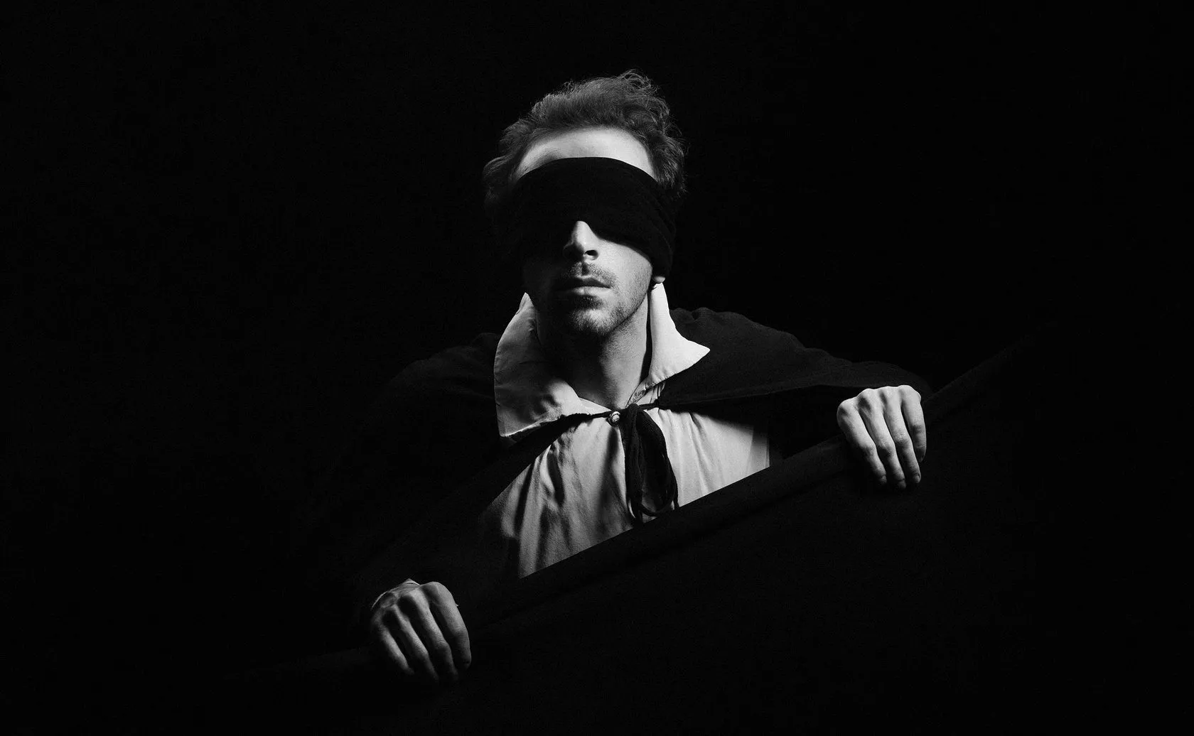 A blindfolded actor in a white shirt and black cape leans on a dark frame.