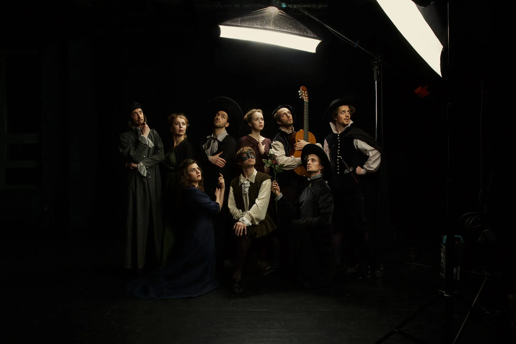 The cast in costume crowd together under a studio light, one holding a guitar.