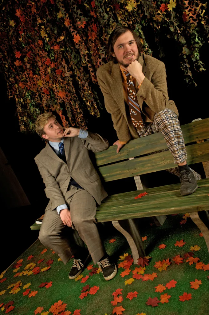 Reference: two men on a park bench in a stage set strewn with autumn leaves.