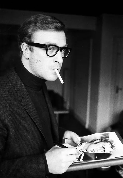 Reference: a man in black-rimmed glasses and a polo neck, a cigarette in his mouth.
