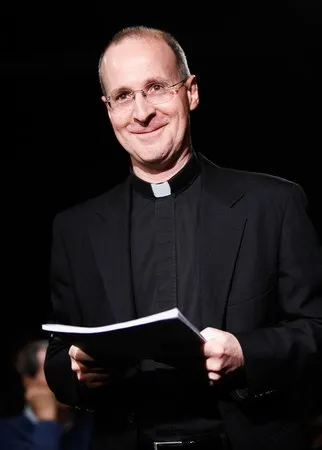 Reference: a smiling man in glasses and a clerical collar holds a script.