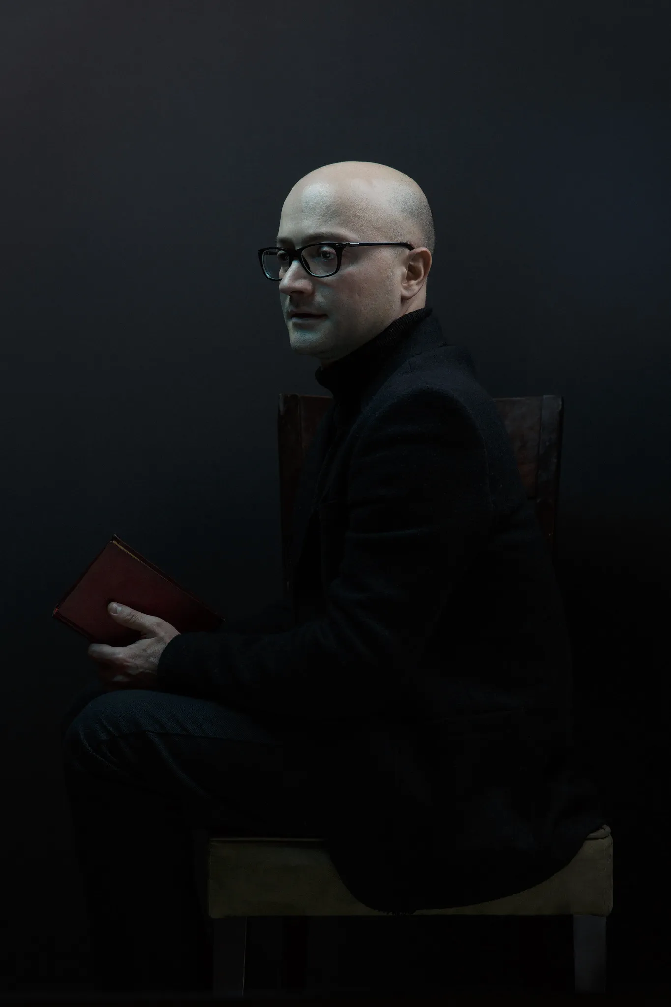 The bald man in glasses sits in profile, a red book on his knee.