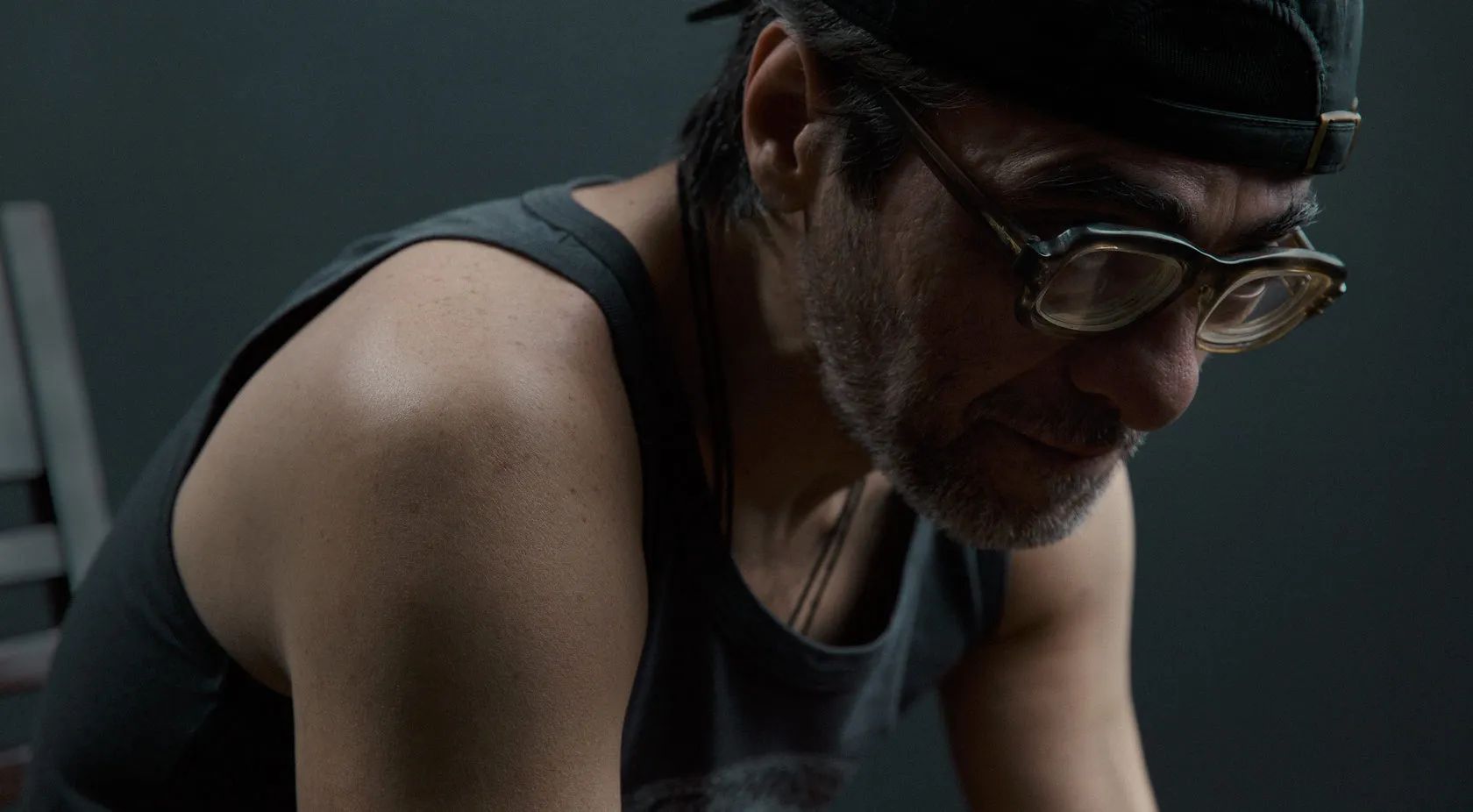 A man in a vest top, cap and glasses leans forward, eyes down.