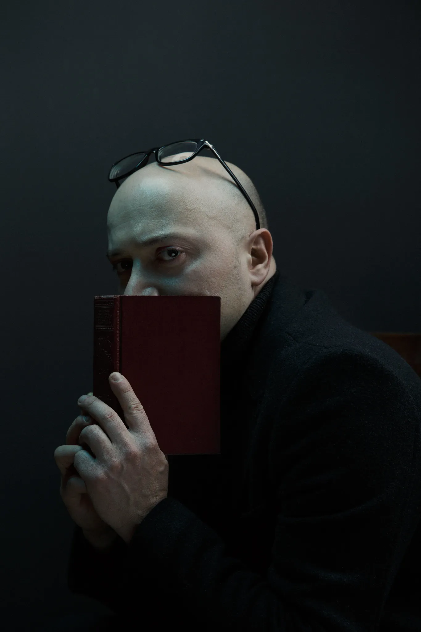 The bald man hides his face behind a red book, glasses pushed up on his head.