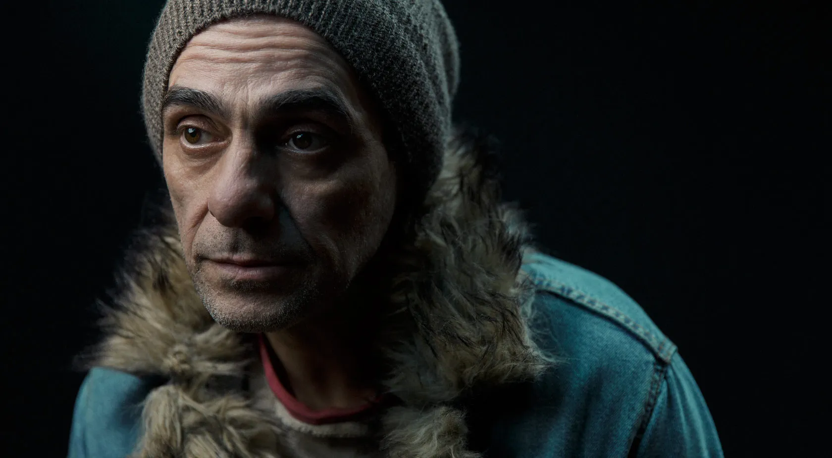 The man in the knit cap and fur-collared denim jacket looks off to the side.