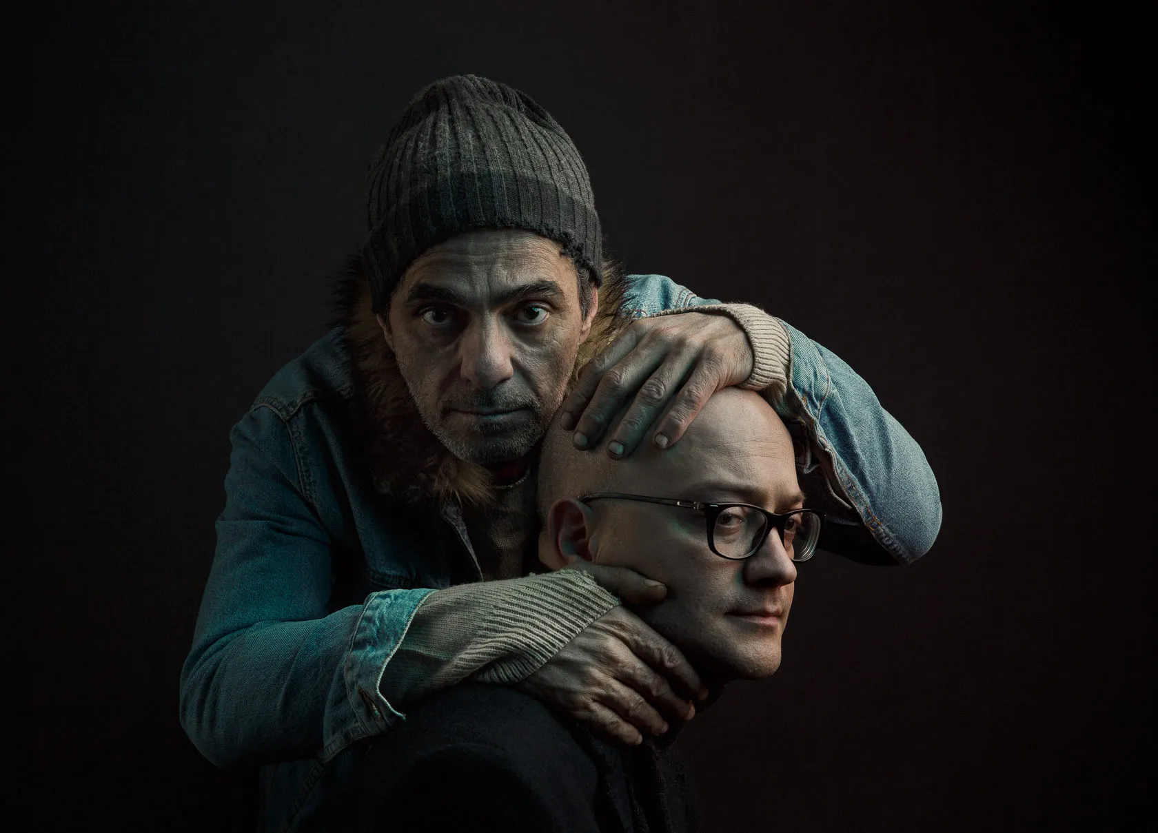 The homeless man in the knit cap holds the bald man’s head against his chest.