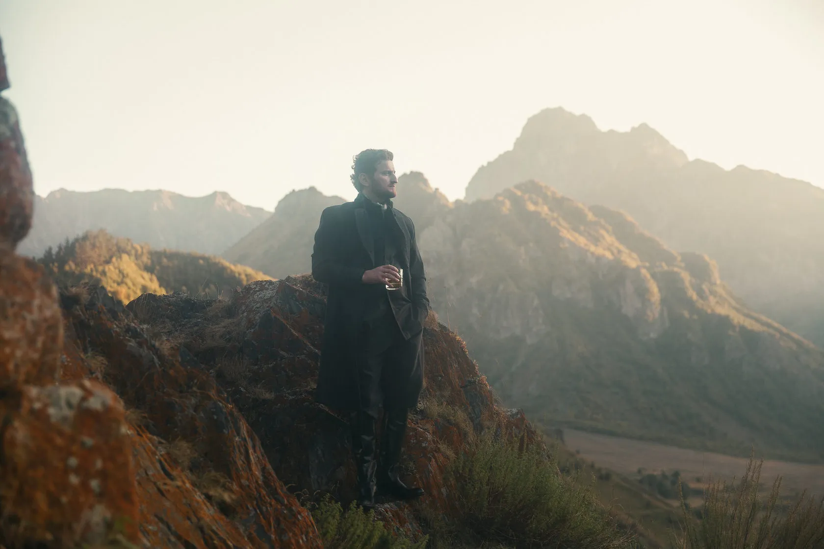 Jimsher Chkhaidze in a long coat stands on a rocky slope, mountains behind him.