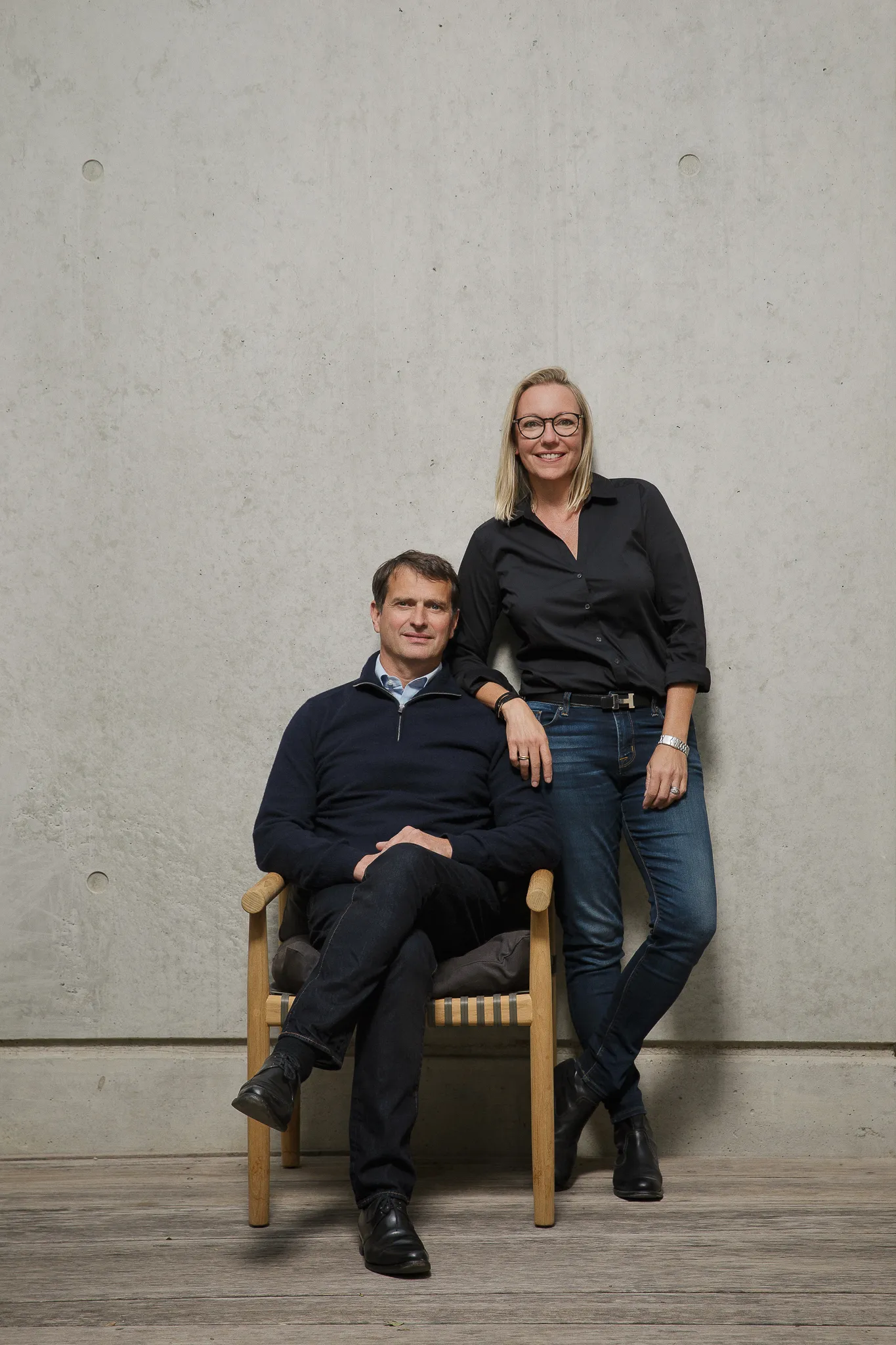 Jean-Louis Chave sits against a concrete wall, a woman in black leaning on his chair.