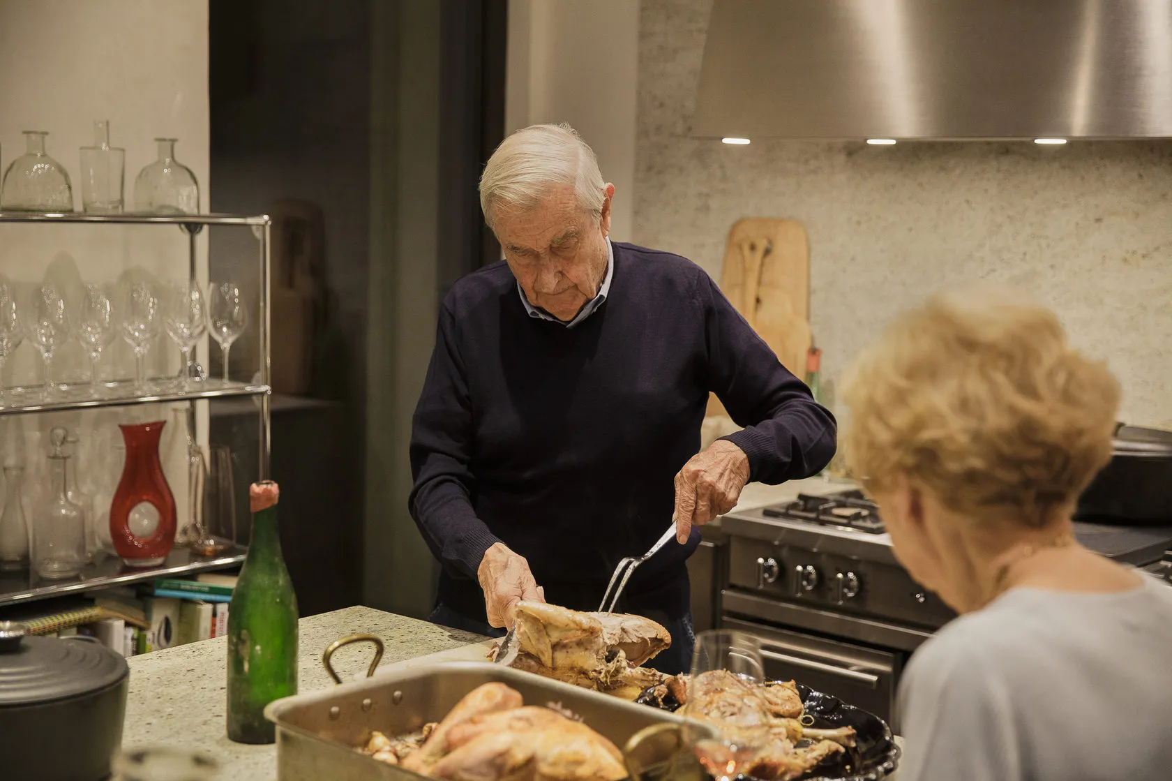 An older man carves a roast in the family kitchen as a woman looks on.