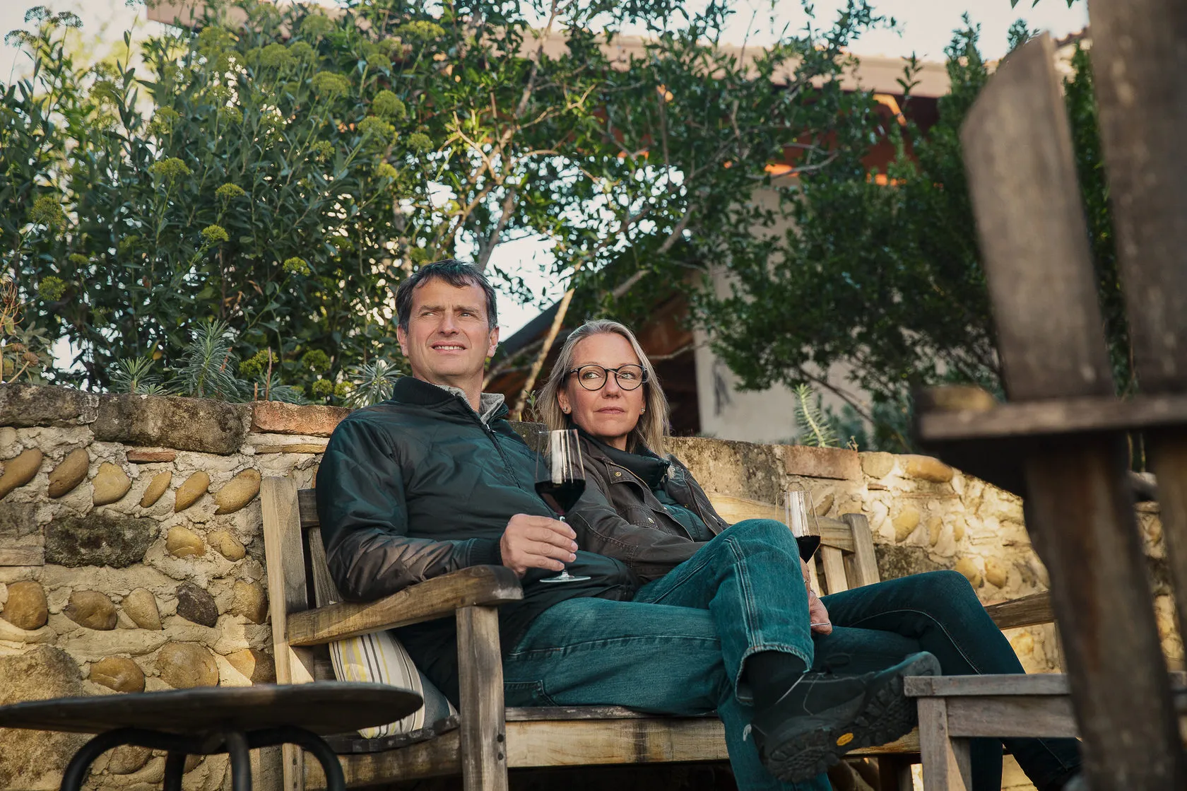 Jean-Louis Chave and a woman sit on a garden bench with glasses of red wine.