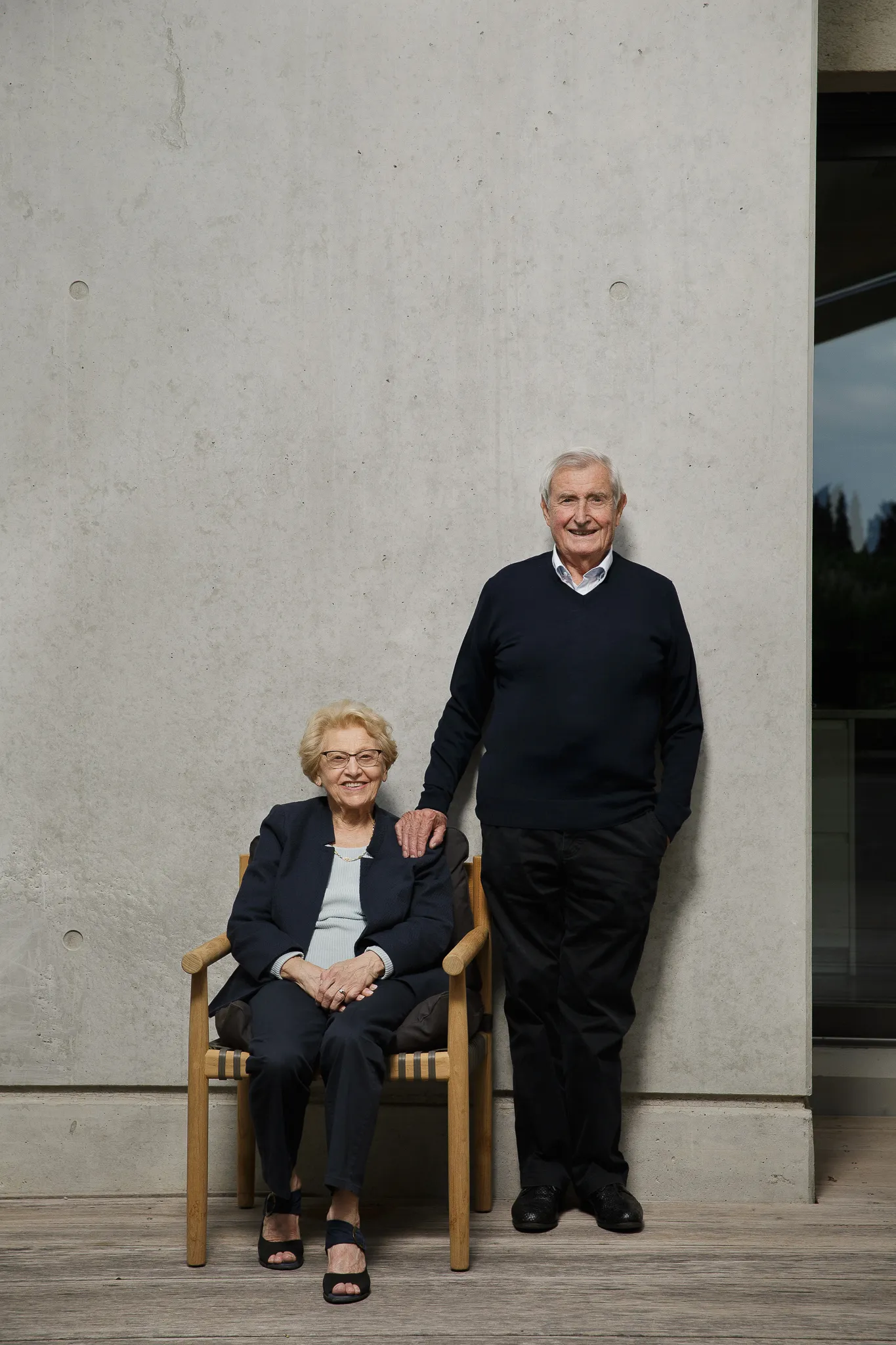 An older couple by a concrete wall, the woman seated, the man standing beside her.