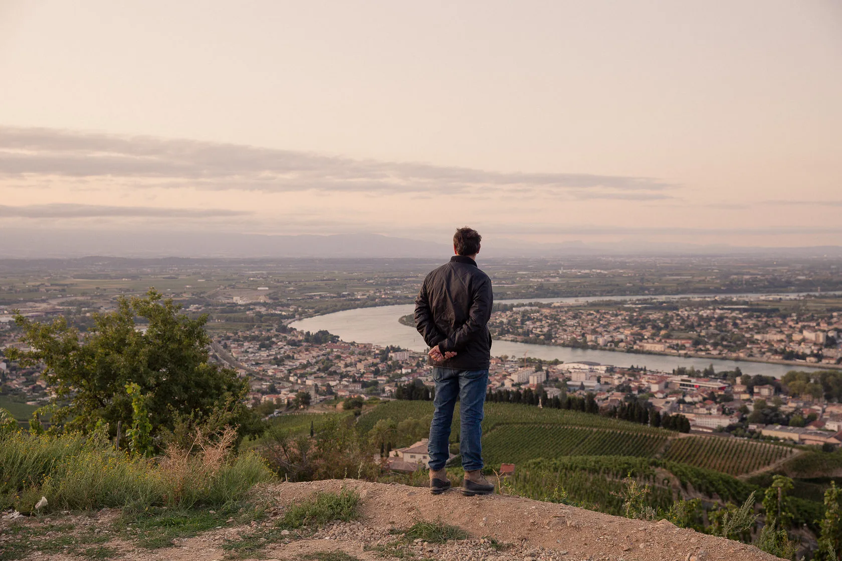 Jean-Louis Chave, seen from behind, looks out over the river and the town at dusk.