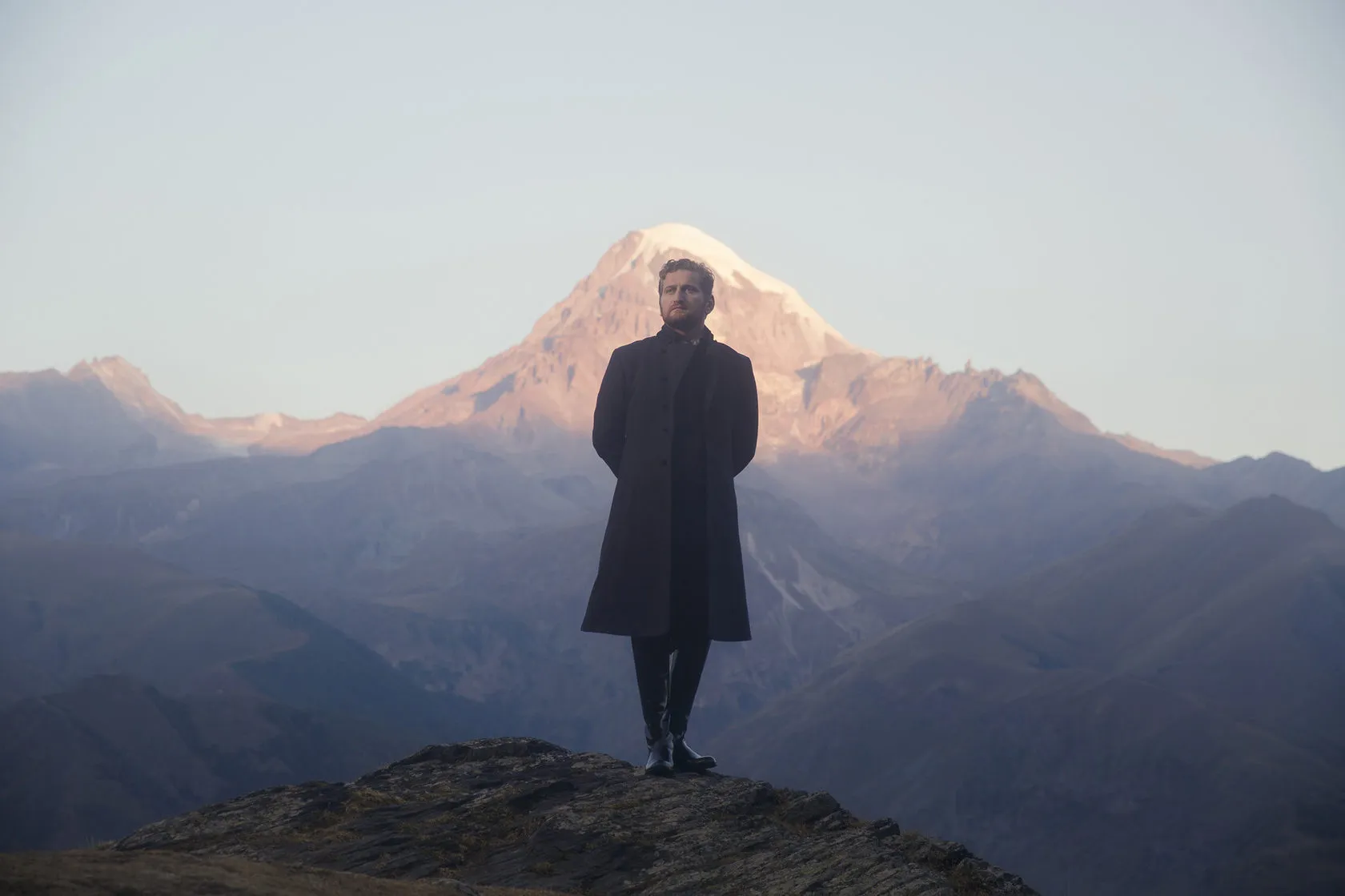 Jimsher Chkhaidze in a long dark coat stands on a ridge below snow-capped Mount Kazbegi.