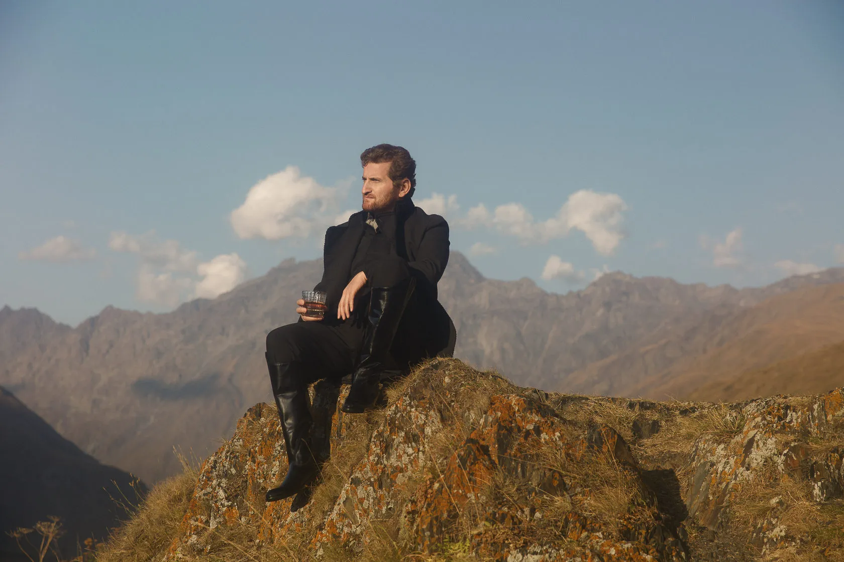 Jimsher Chkhaidze sits on a rocky outcrop with a glass of whisky, mountains and clouds behind.