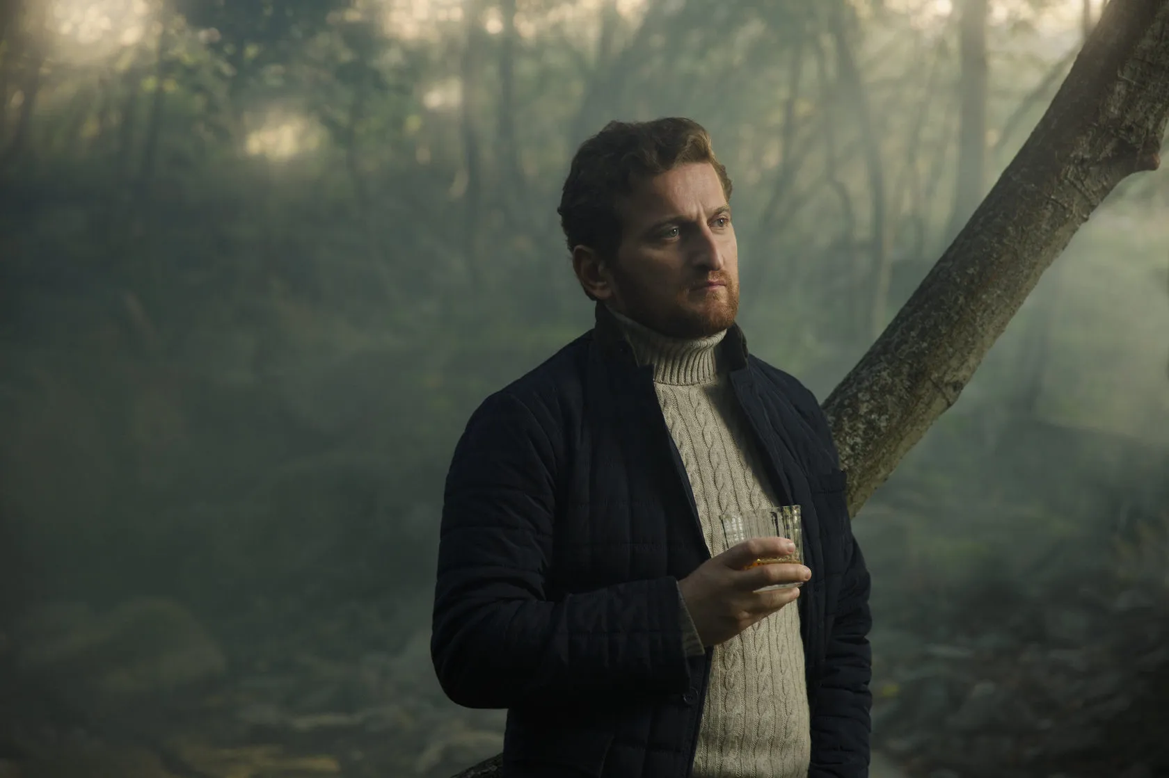 Jimsher Chkhaidze in a cable-knit jumper holds a glass in a misty forest.
