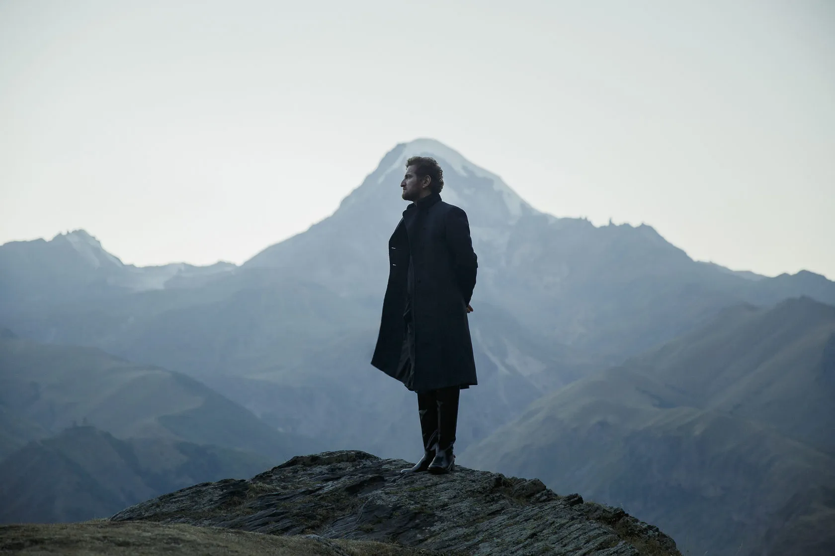 Jimsher Chkhaidze in a long coat stands in profile on a rock, blue mountains behind.
