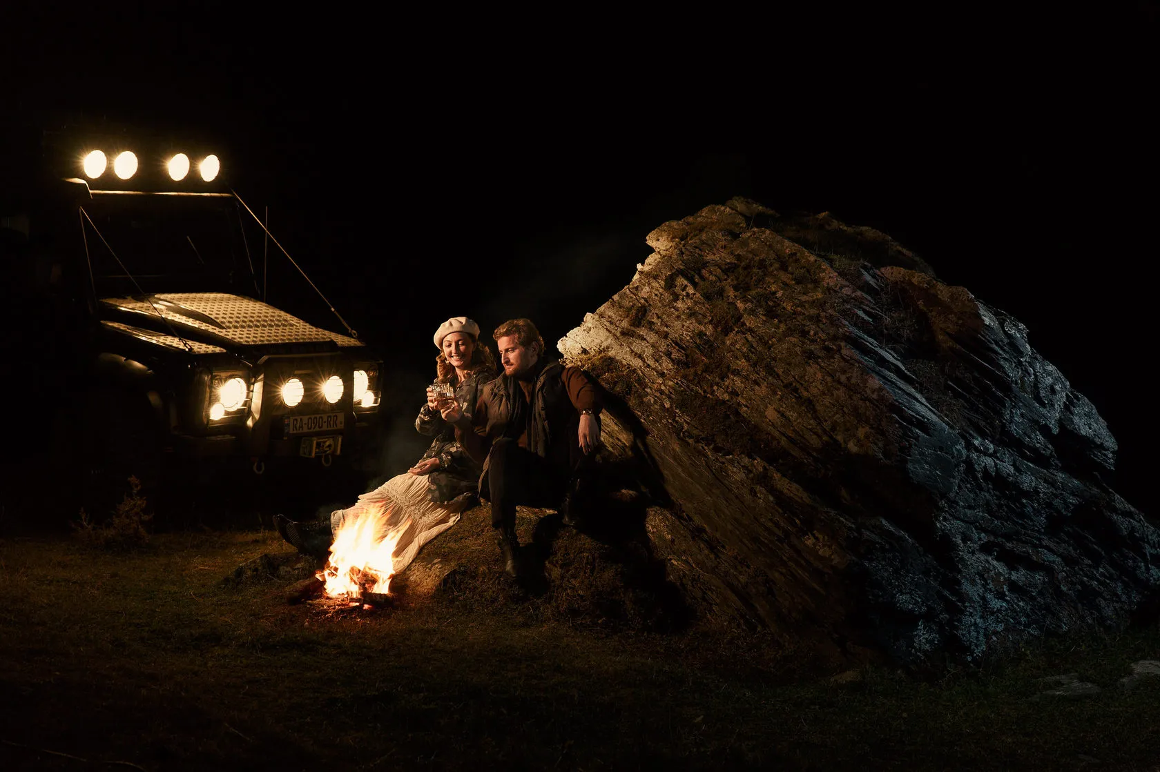 Two people sit by a campfire against a boulder at night, a truck’s lights glowing behind.