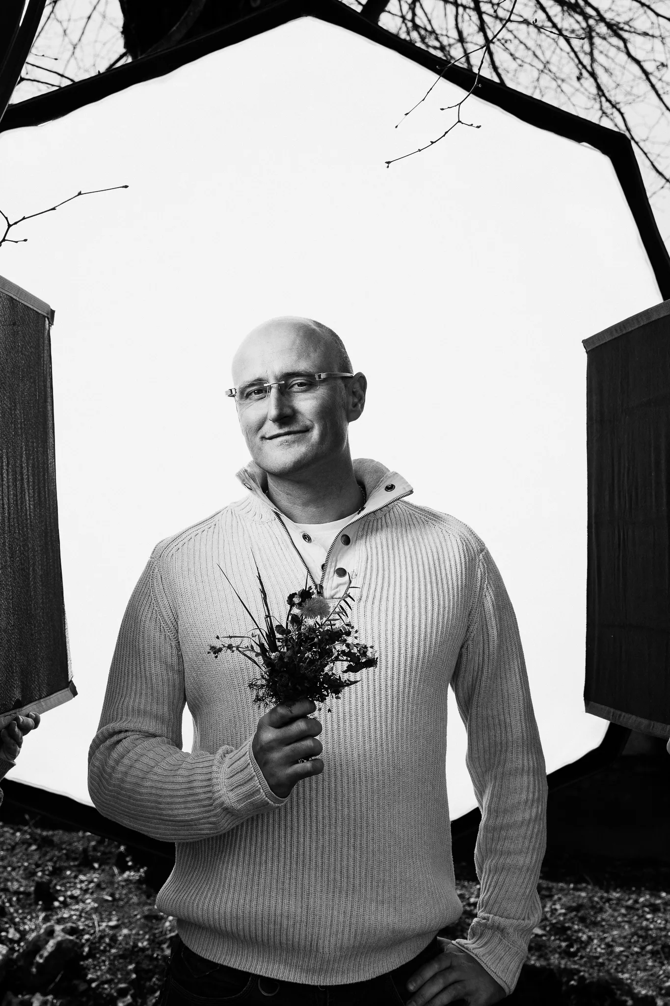 A bald winemaker in a knit jumper holds a posy of wild flowers outdoors.