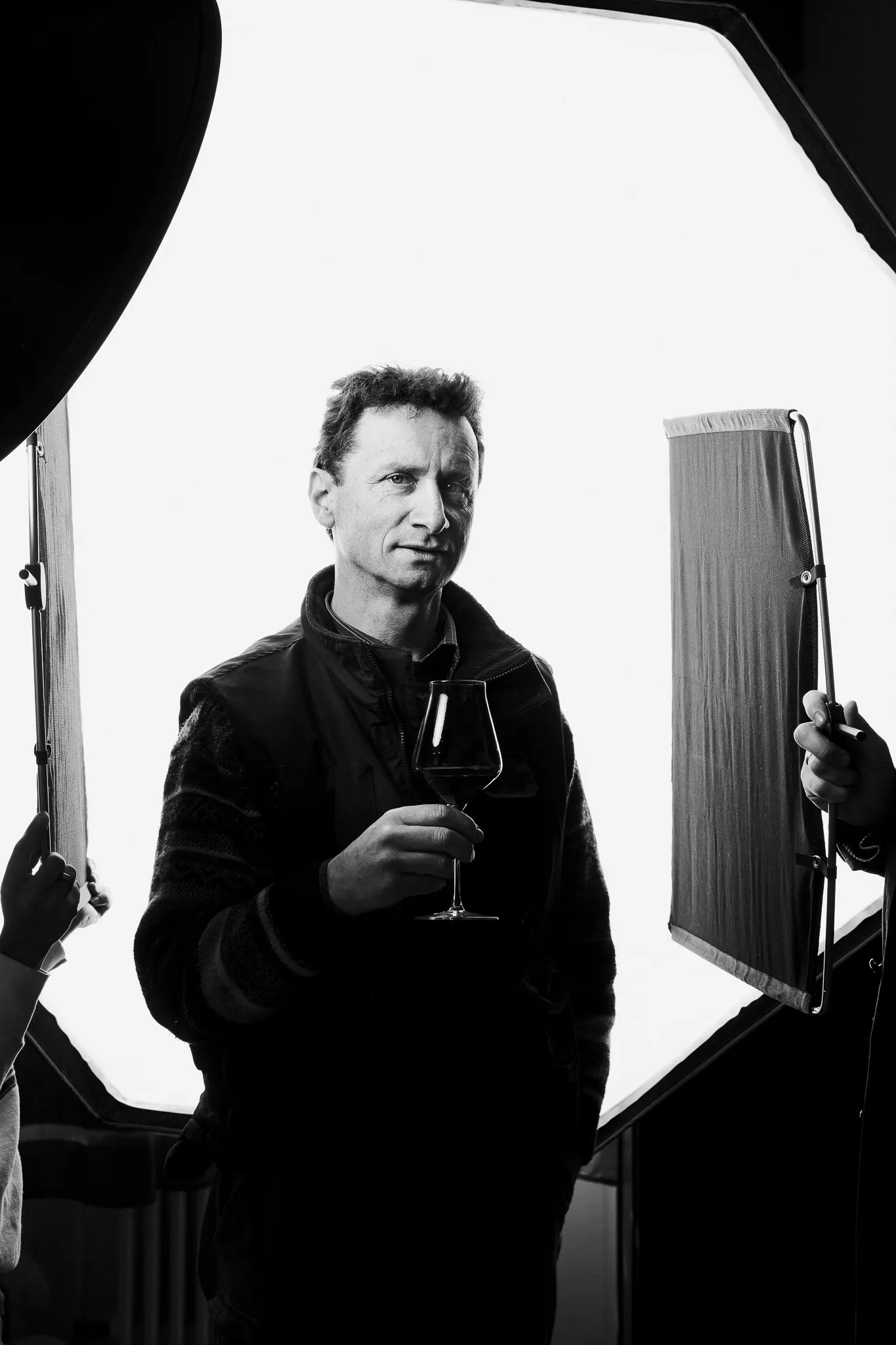 A winemaker holds a glass of red wine between studio lights.