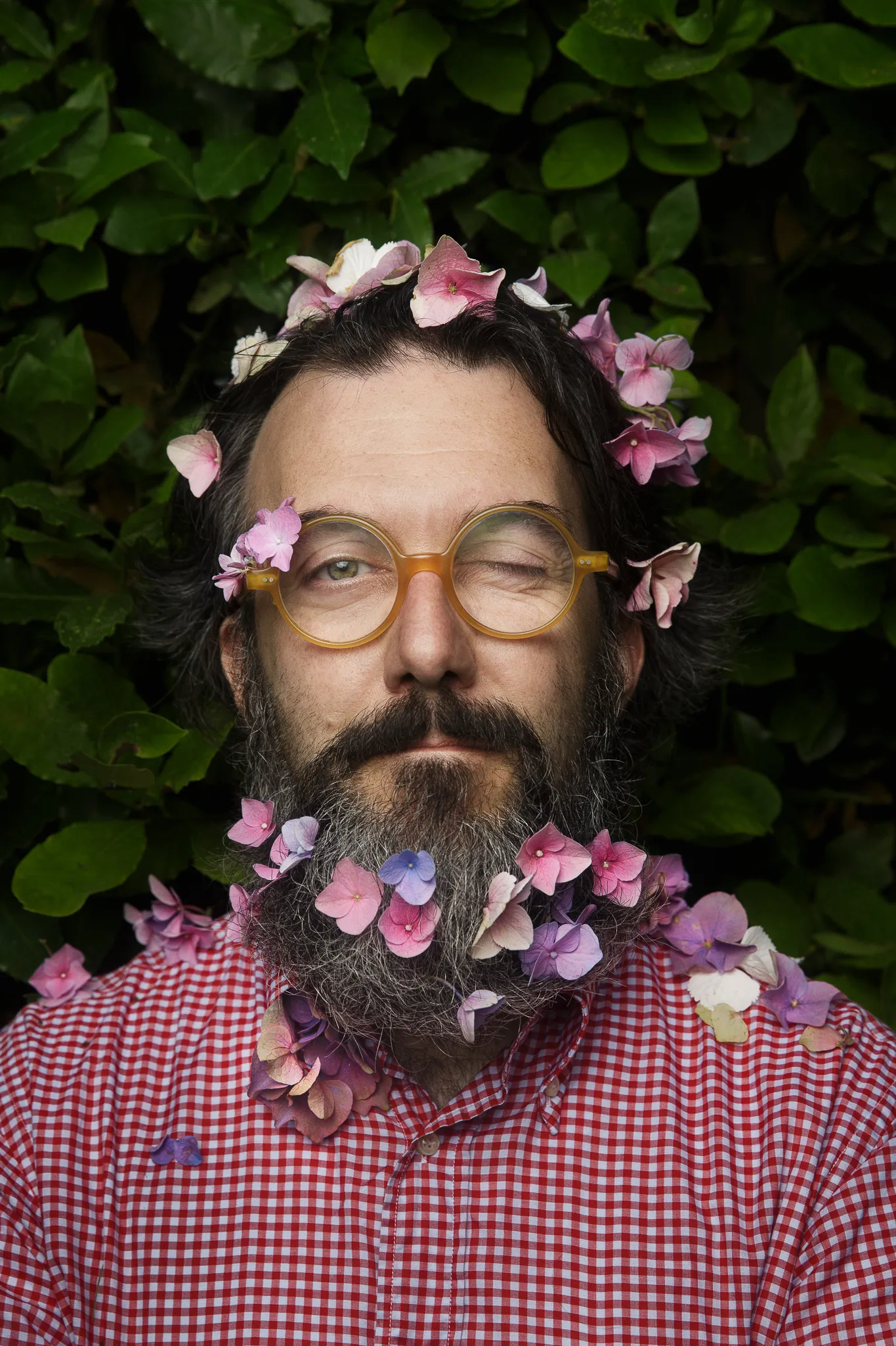 Marco Casolanetti, flowers in his hair and beard, against a hedge of leaves.