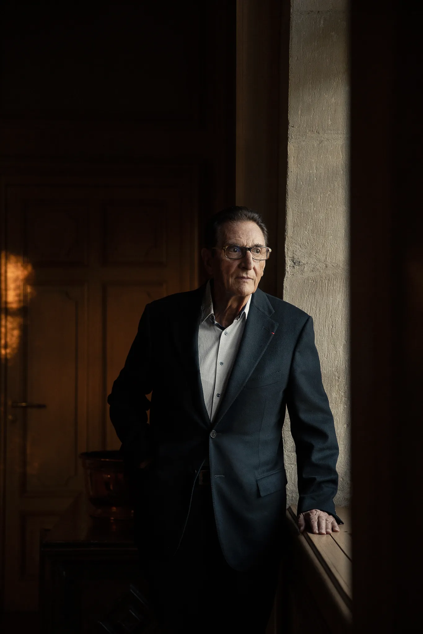 Marcel Guigal in a dark suit stands by a tall window in a dim room.