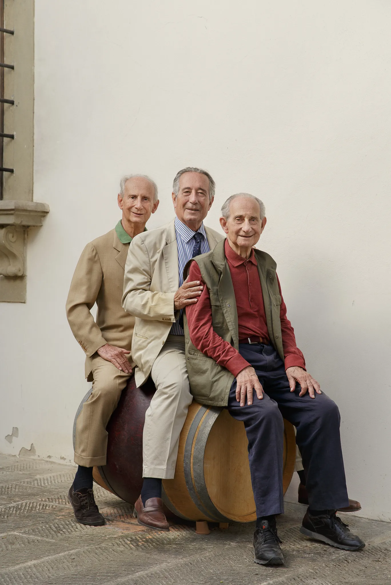 Three Frescobaldi men in jackets sit on a wine barrel against a pale wall.