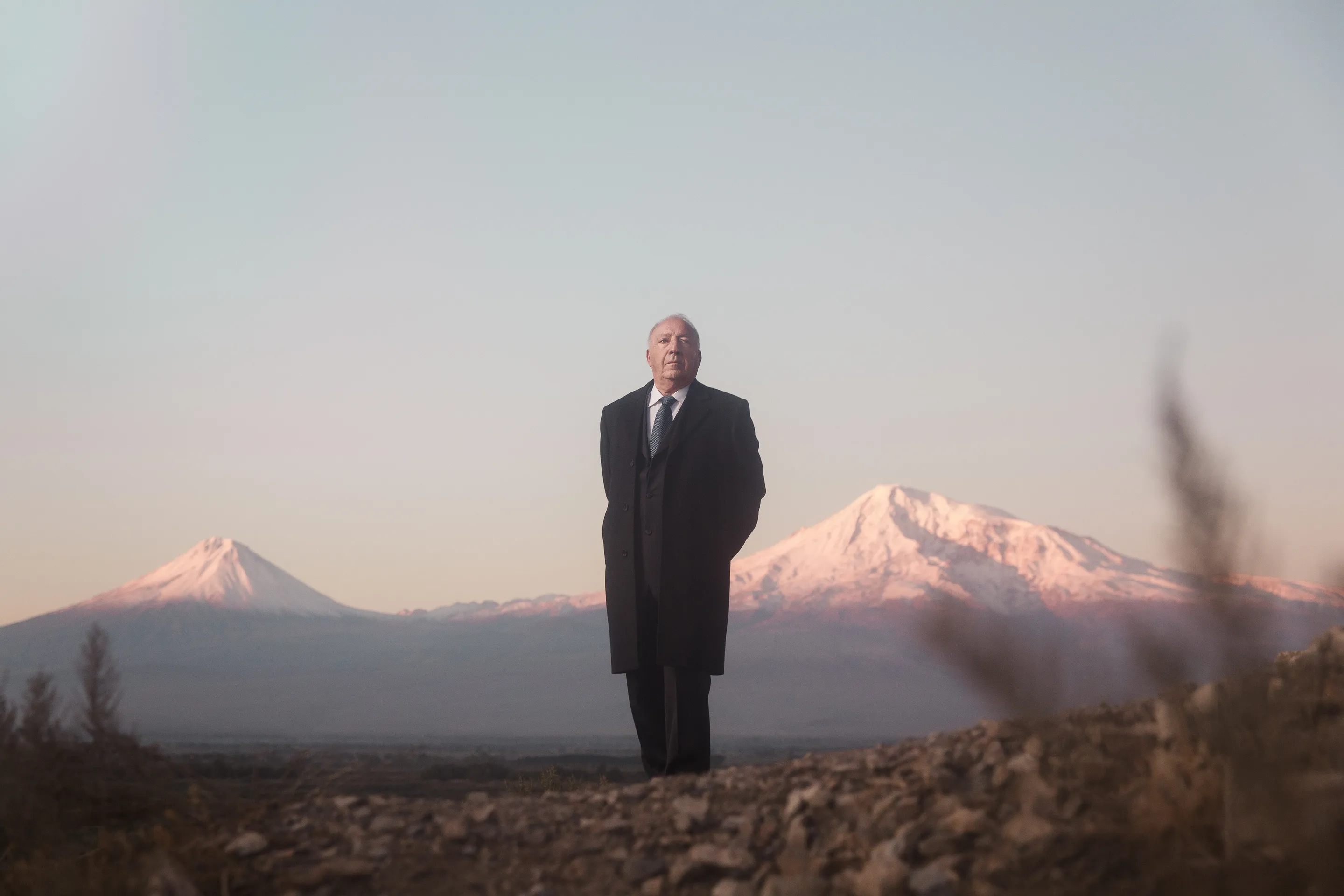 Simple Wine News portraits by Andrei Kovalev: a man in a dark suit stands alone in a field between two snow-capped twin peaks.