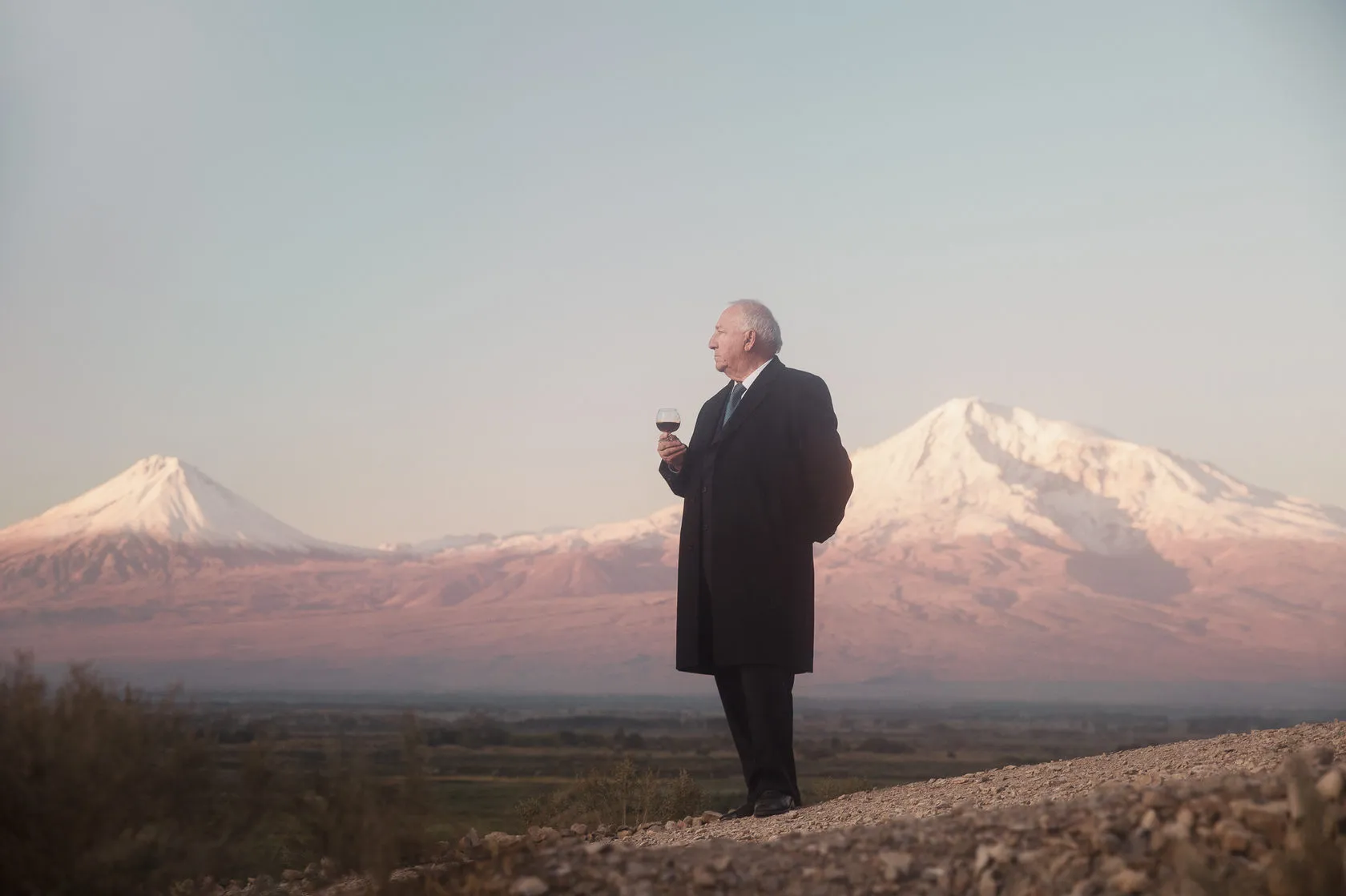 Robert Azaryan in a long dark coat holds a glass of wine, snowy twin peaks behind him.