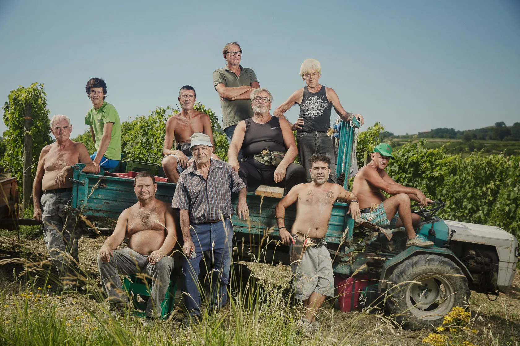 Walter Massa’s team, some shirtless, crowd onto a tractor and trailer in a vineyard.