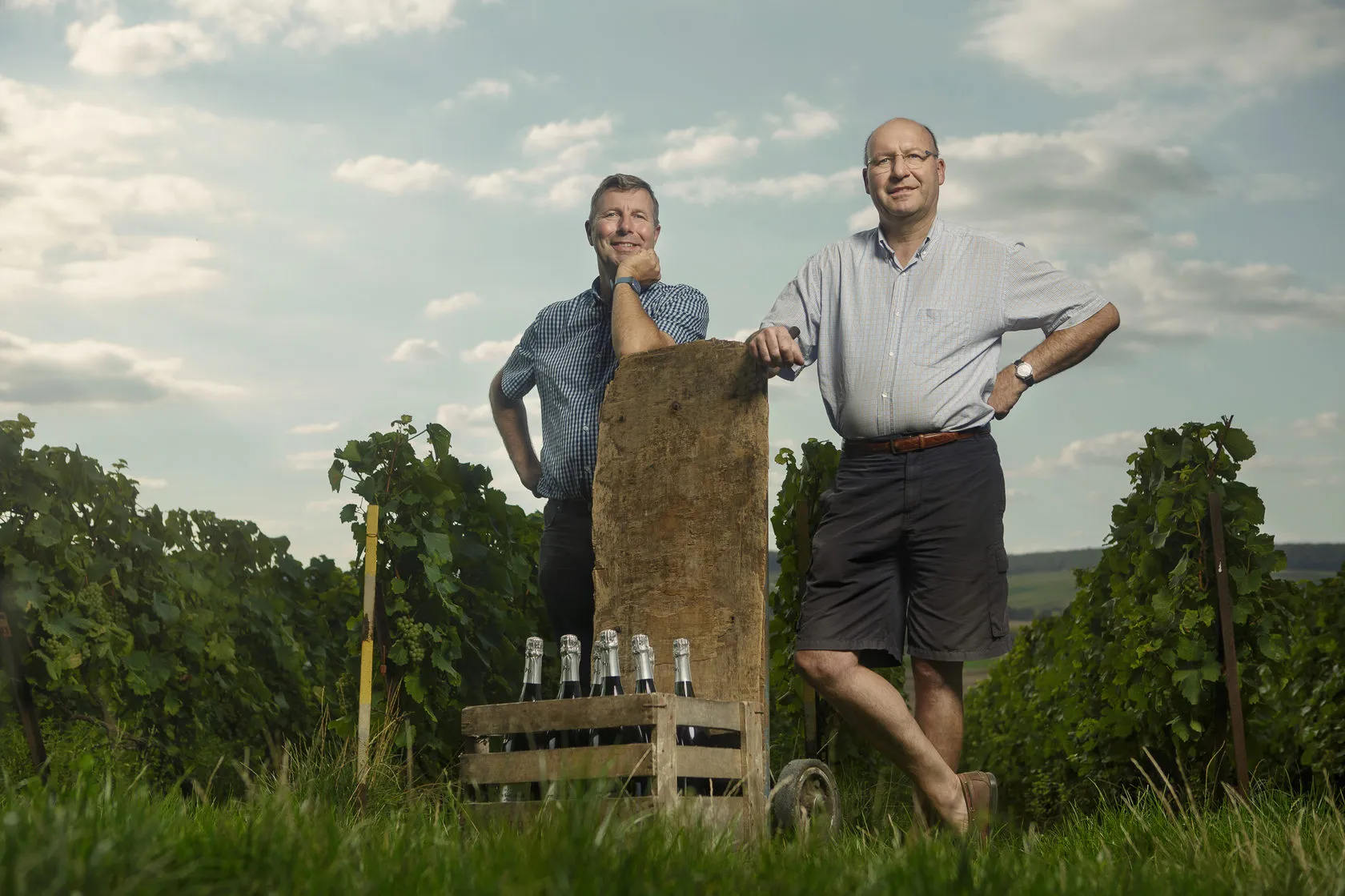 Didier and Olivier Gimonnet in a vineyard, an old stone post and a crate of bottles between them.