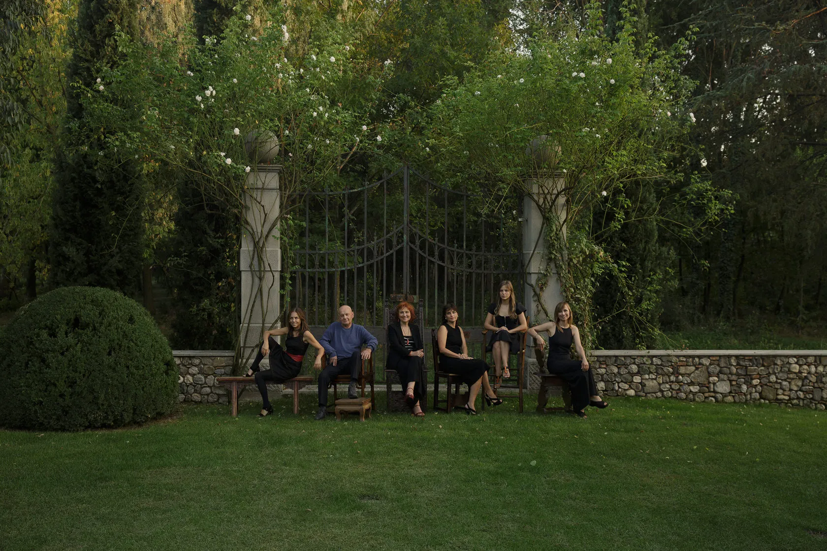The Nonino family sits on a bench in front of tall iron gates in a garden.