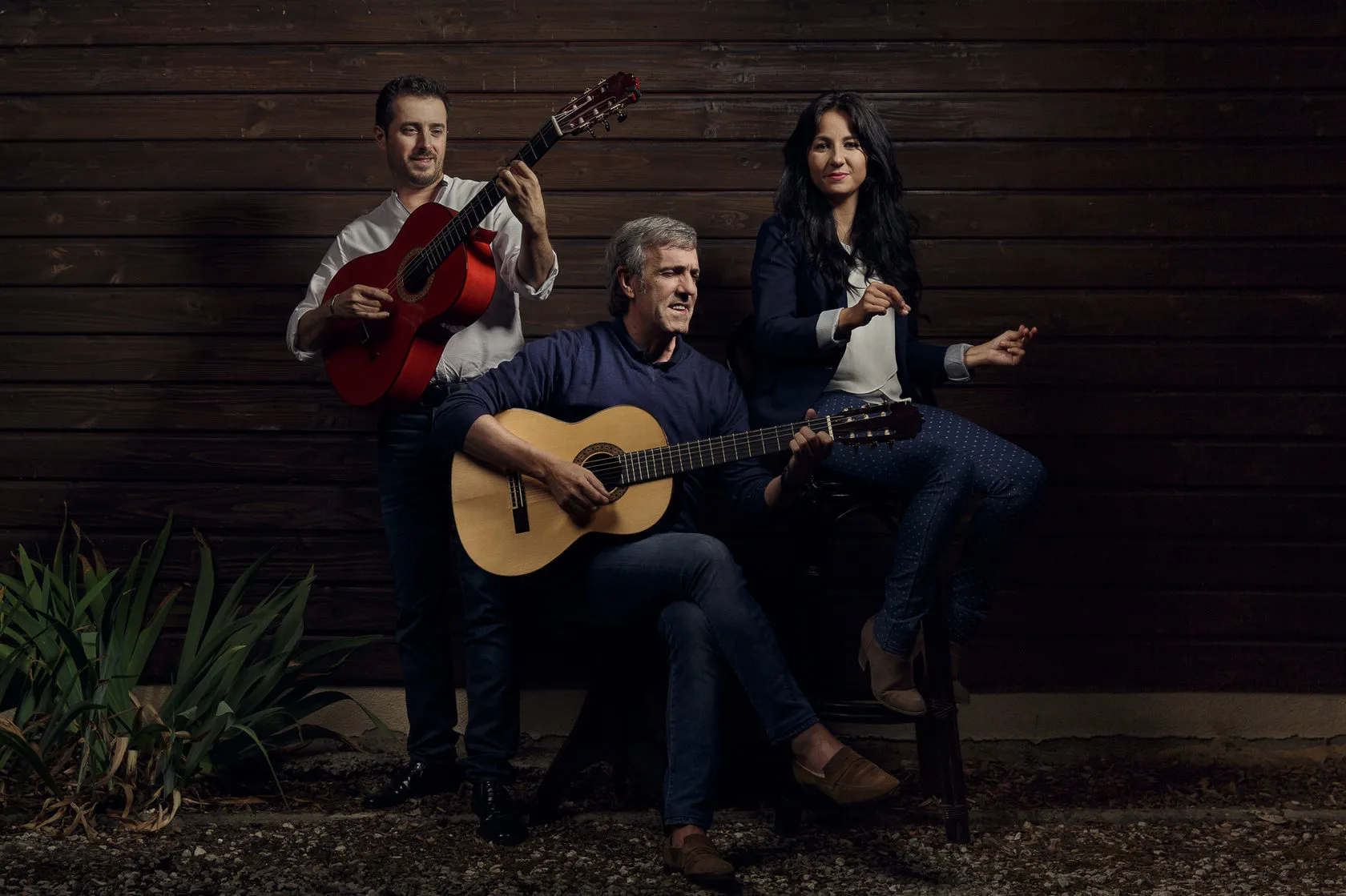 José Moro and two others play guitar and clap against a dark wooden wall.