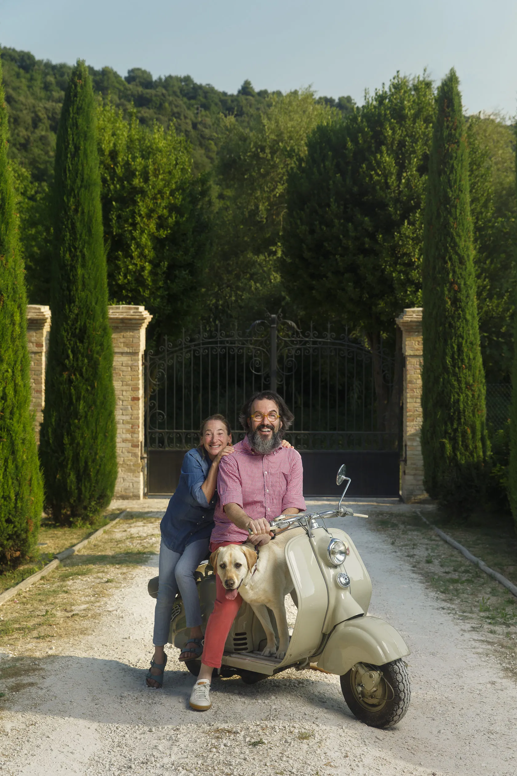 Eleonora Rossi and Marco Casolanetti on a vintage scooter with a dog, by iron gates and cypresses.