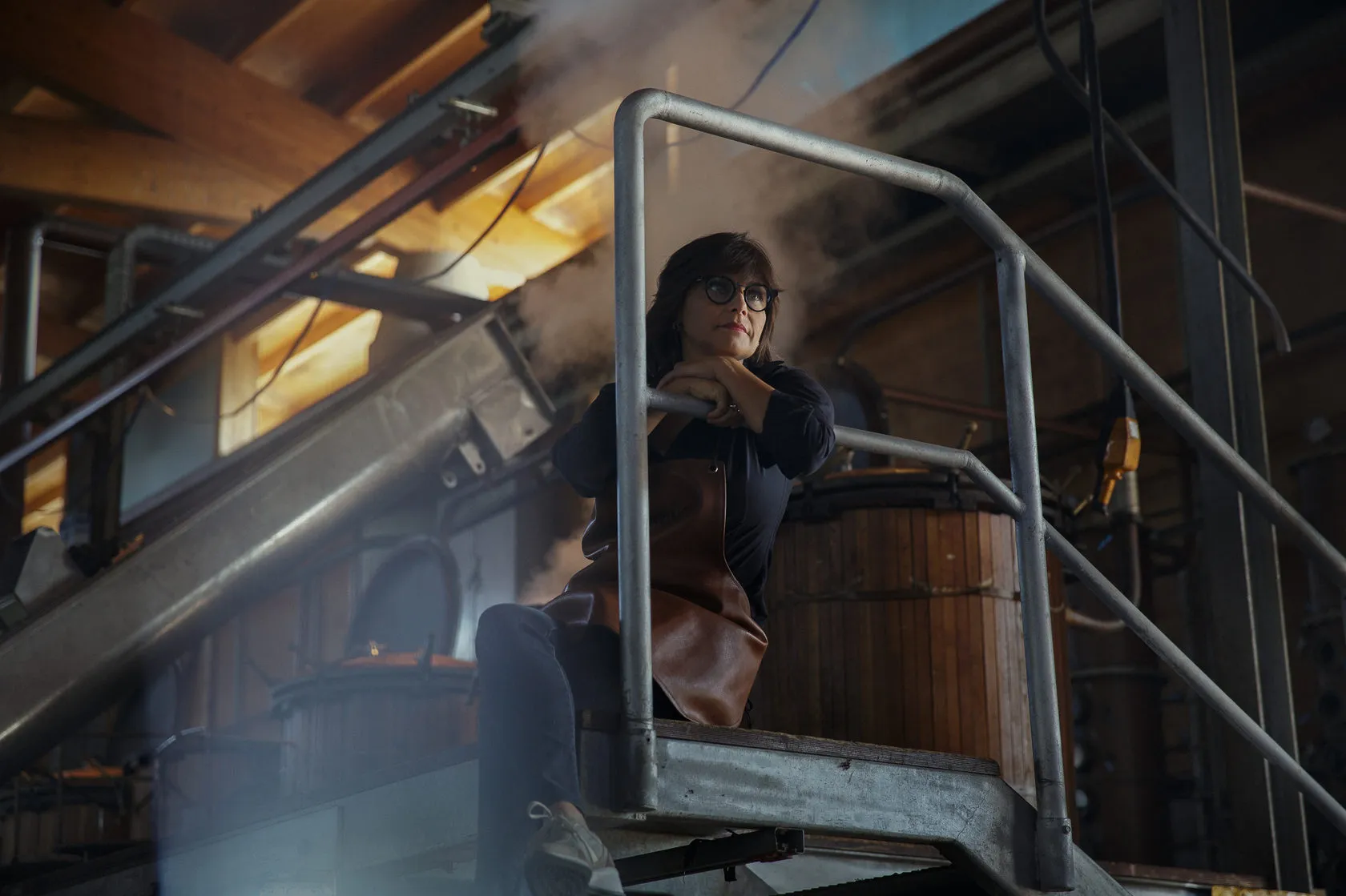 A woman in a leather apron sits on a steel walkway in the steamy distillery.