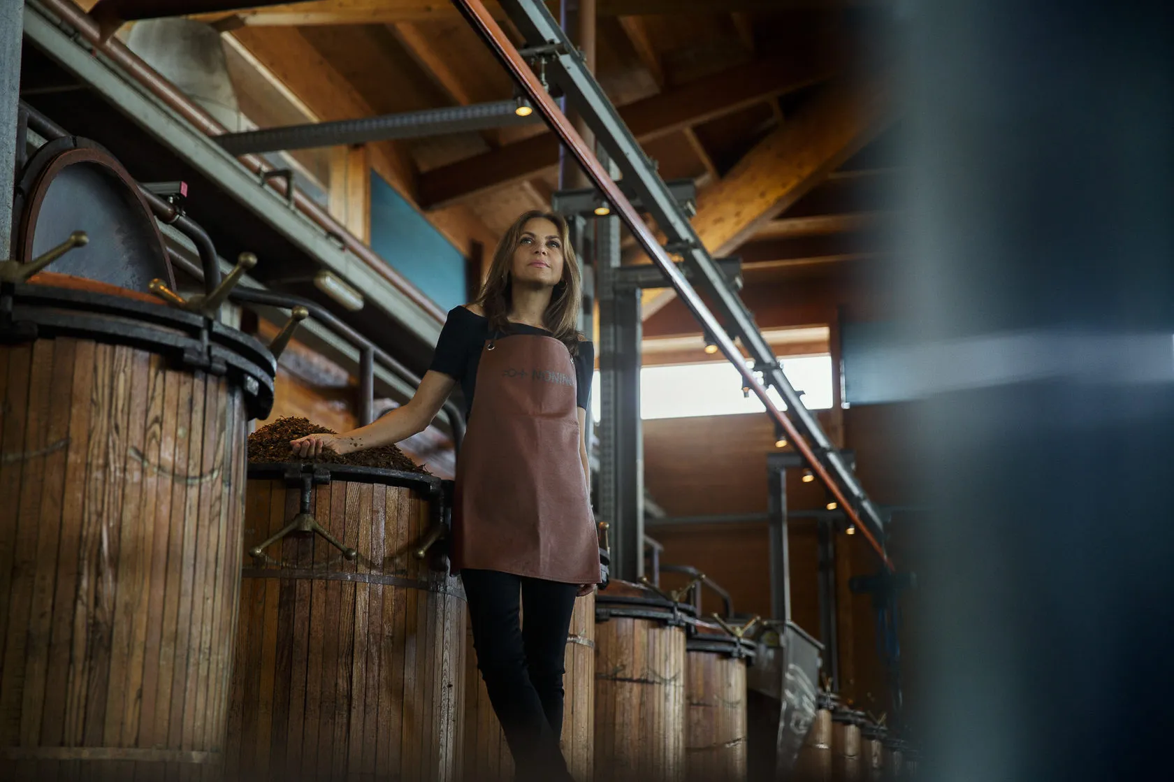 A woman in a leather apron leans on a wooden still under a timber roof.