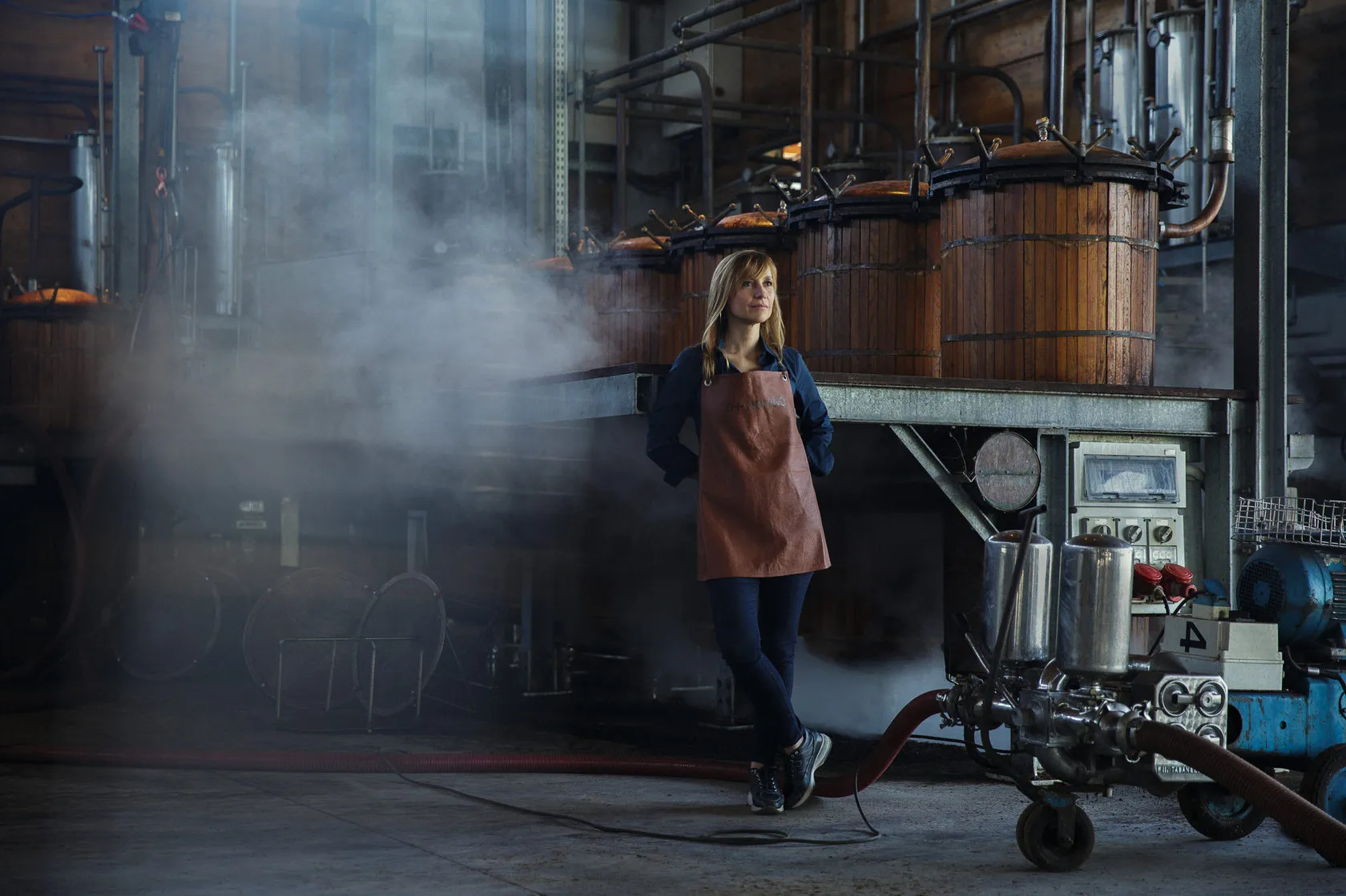 A woman in a leather apron stands in the steam beside the stills.