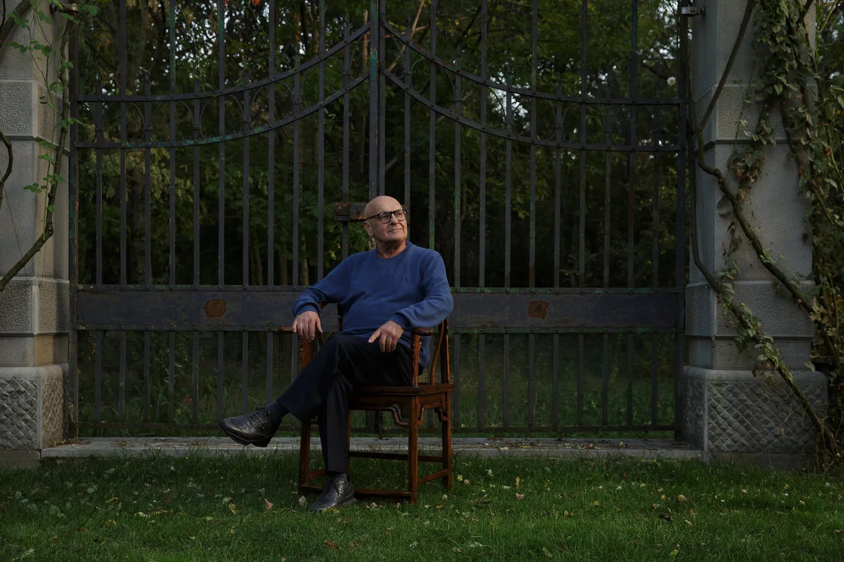 An older man in a blue jumper sits on a chair in front of tall iron gates.