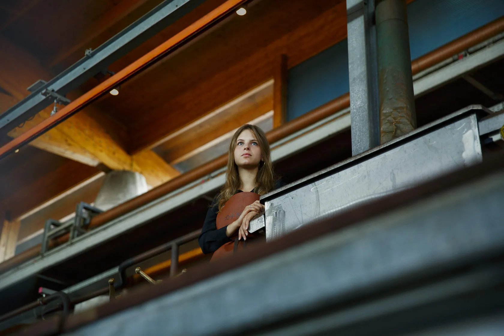 A young woman leans on a metal rail in the distillery, looking up.