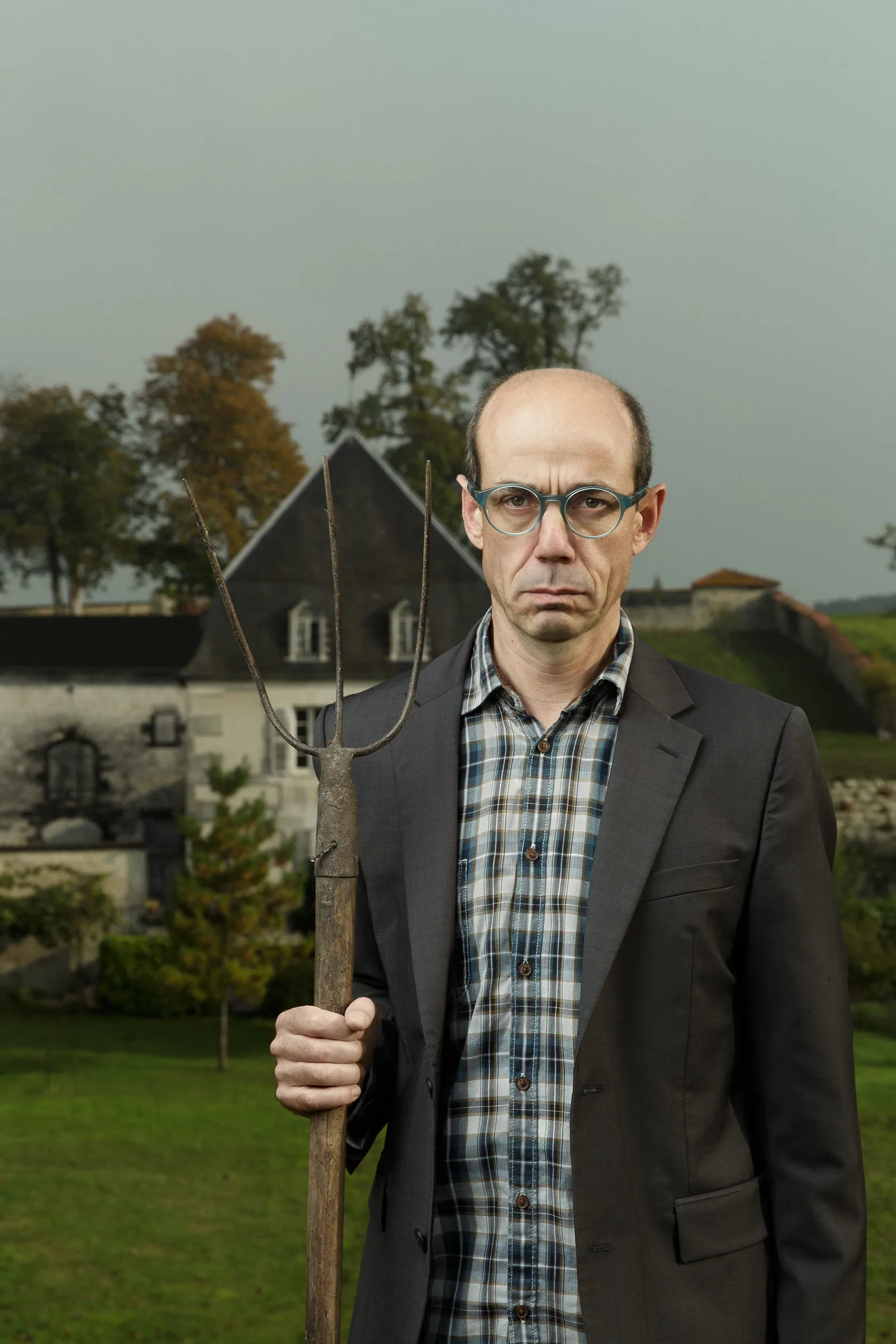 A bald winemaker in glasses holds a pitchfork in front of an old house.