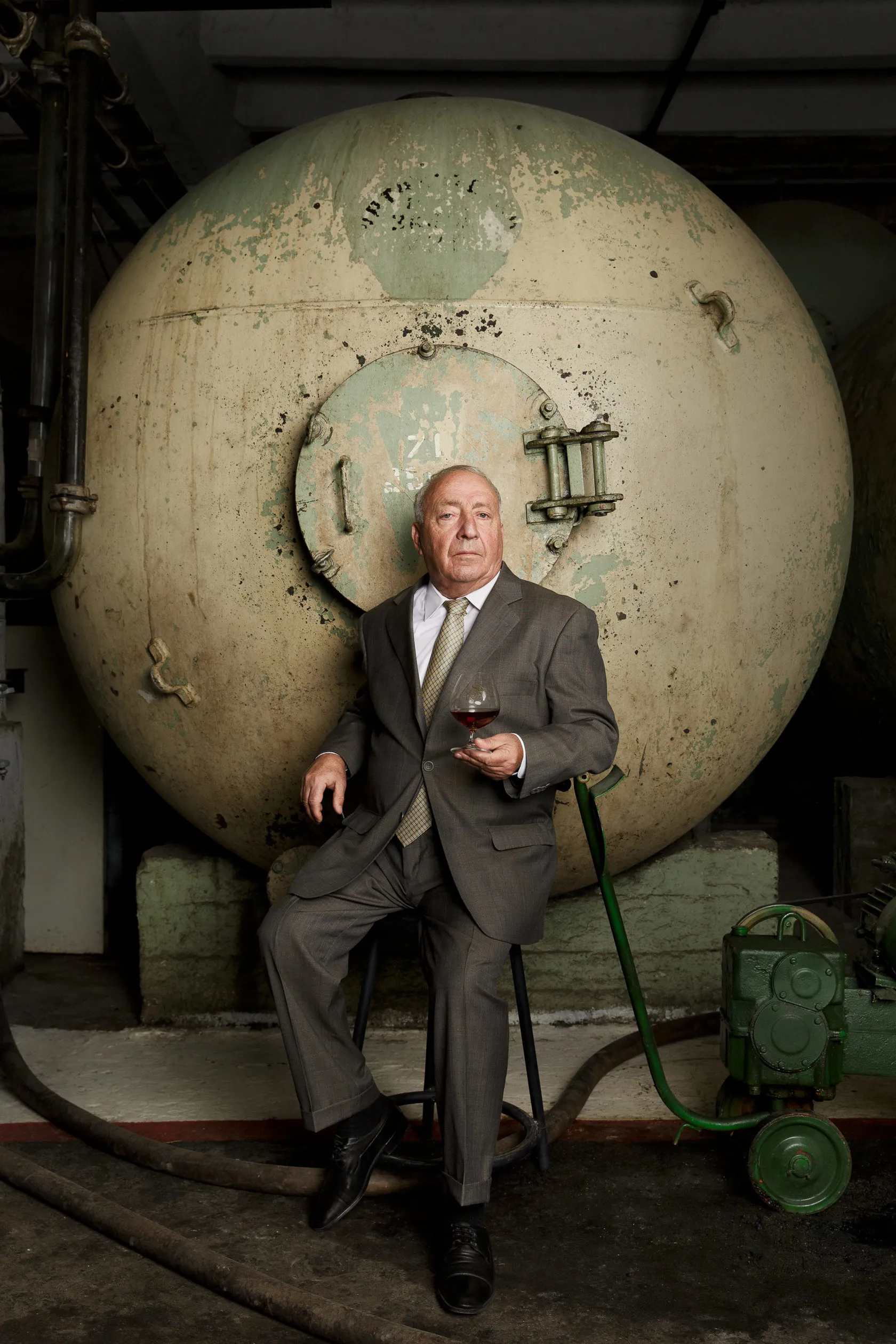 An older winemaker in a suit sits with a glass in front of a huge round tank.