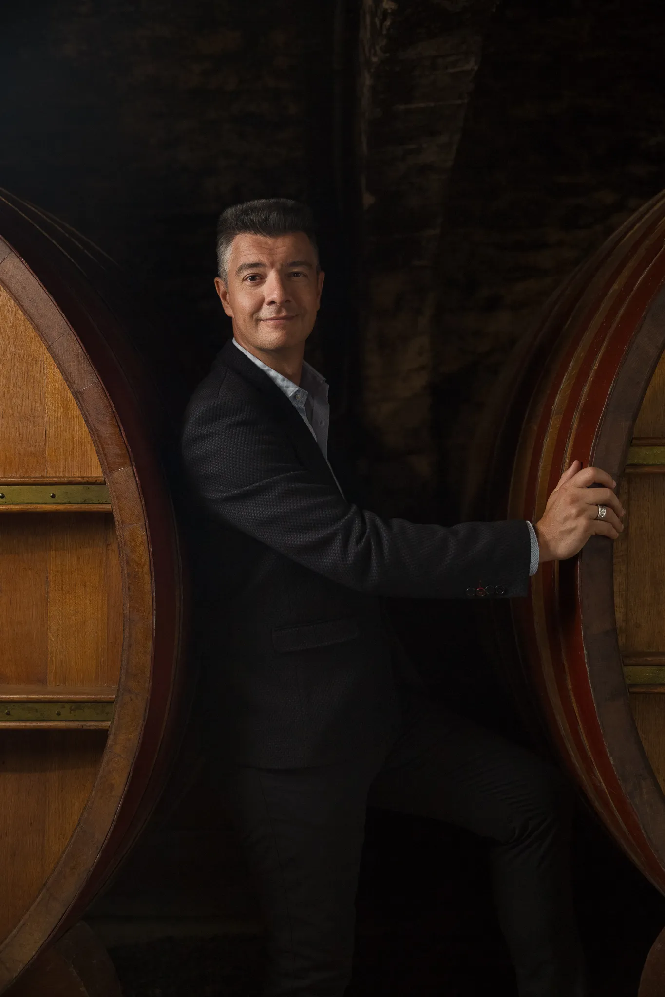 A winemaker in a dark suit stands between two great oak casks.